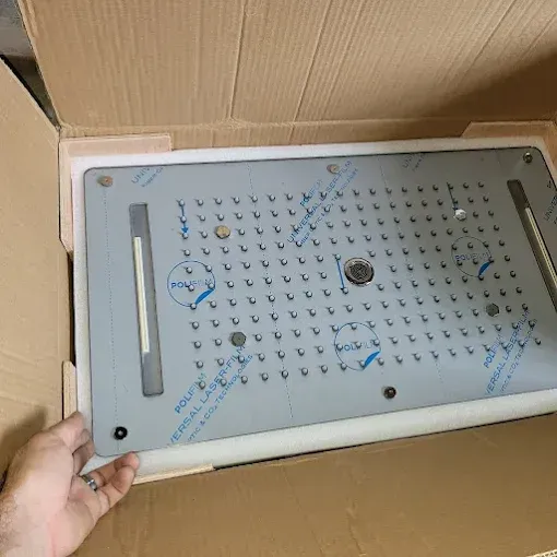 A person holds a large, rectangular stainless steel rainfall showerhead with protective film inside a cardboard box.