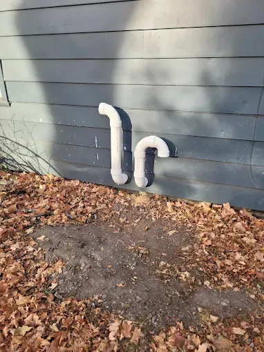 Two white PVC pipes emerge from grey house siding and curve downward, positioned above a ground covered in dry leaves.