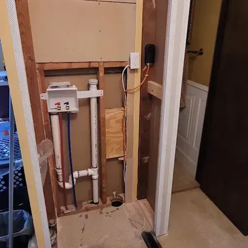 Wall cavity exposing white PVC plumbing for a laundry hookup, electrical outlet, and wiring against unfinished drywall.