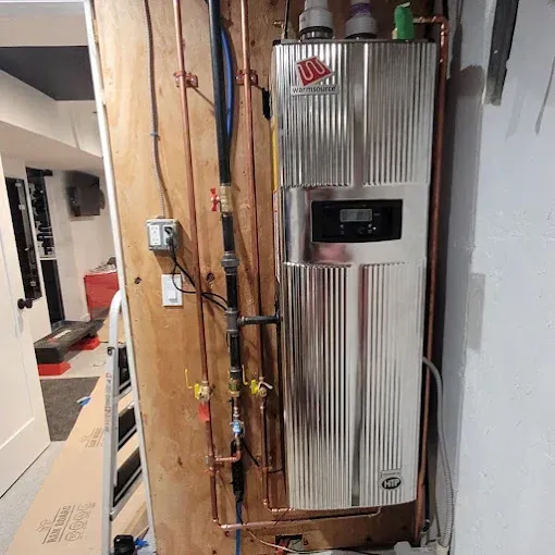 A shiny, silver wall-mounted water heater with copper piping and controls installed on a wooden panel in a utility room.