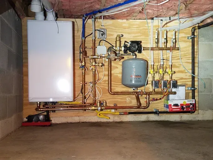 A wall-mounted residential heating system with a white boiler, copper piping, and an expansion tank on a wooden board.