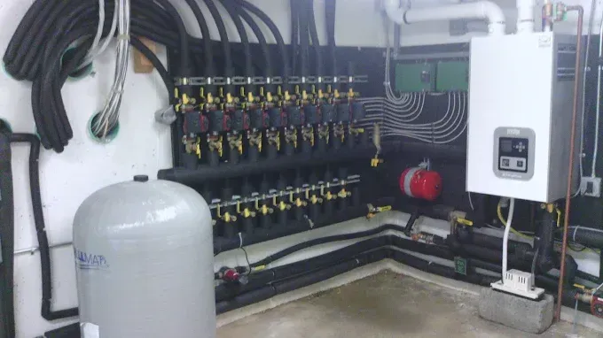 A mechanical room featuring a white wall-mounted boiler, a large gray expansion tank, and a multi-zone piping manifold.