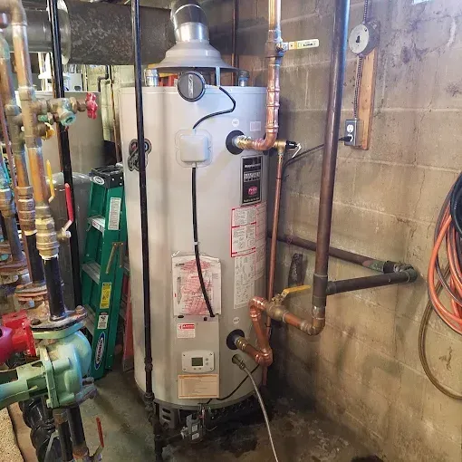 A gray gas water heater stands in a basement with various copper pipes, valves, and a nearby green stepladder.