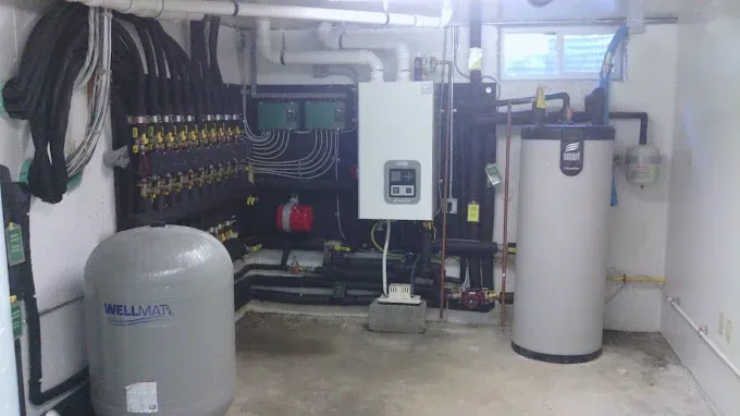 A mechanical room with a white boiler, a tall gray hot water tank, a large expansion tank, and a radiant heating manifold.