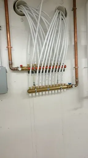 Two brass plumbing manifolds mounted on a white wall, connected to copper pipes and multiple white PEX water lines.