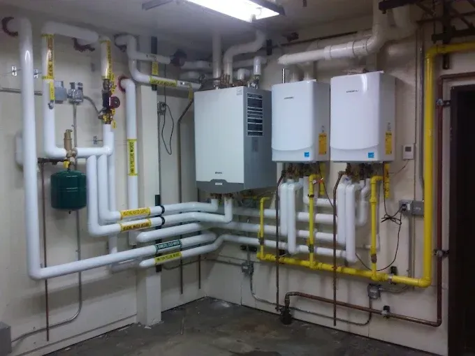 A mechanical room with white, wall-mounted boilers connected by extensive white piping and yellow gas lines on a wall.