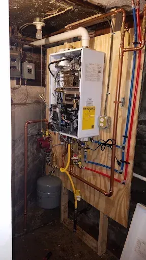 A wall-mounted tankless water heater with copper piping and exposed wiring installed on a wooden board in a basement.