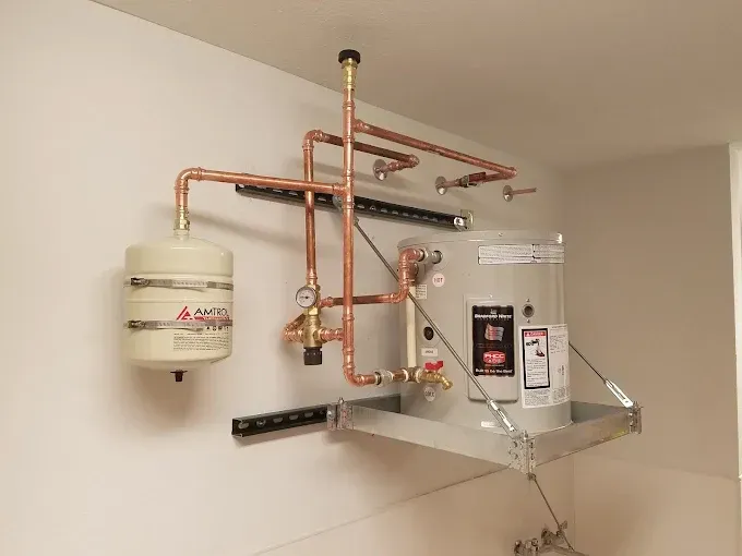 A wall-mounted electric water heater with an expansion tank, copper piping, and safety valves on a metal shelf.