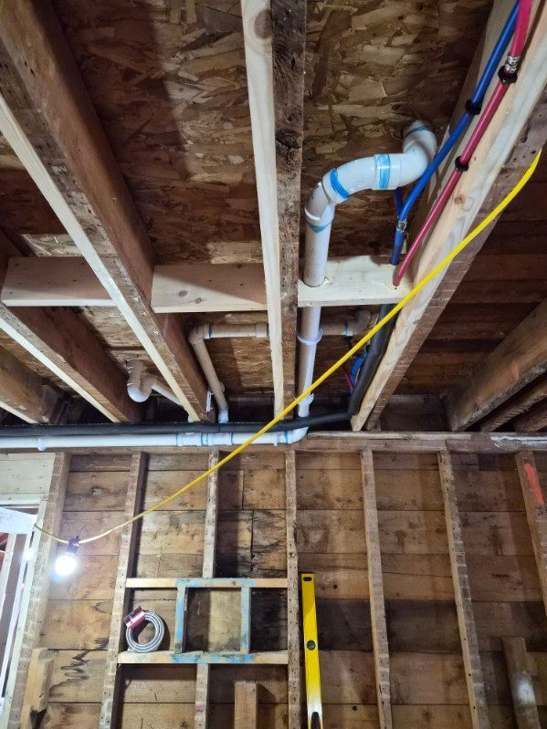 An unfinished basement ceiling showing exposed floor joists, white PVC plumbing pipes, and red and blue PEX water lines.