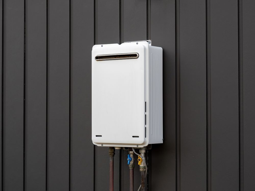 A white tankless water heater mounted on a dark, vertical-paneled outdoor wall with visible utility pipes below.