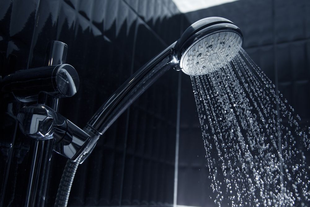 A close-up of a modern silver shower head spraying water against a dark, textured tiled background.