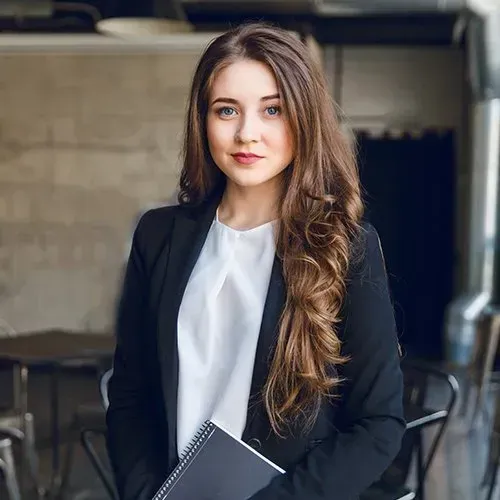 Woman in a black blazer and white blouse holding a notebook, standing in an office.