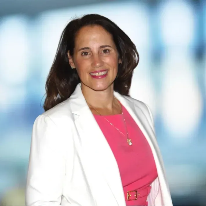 Woman with dark hair in white blazer, pink dress, smiling against blurred background.