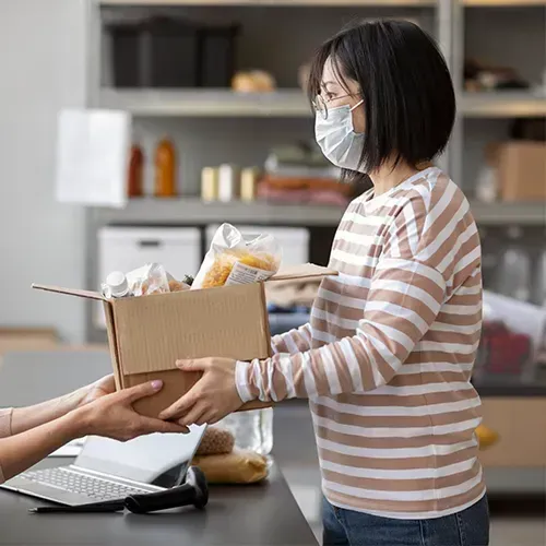 Woman in mask hands a box of food to another person at a food bank.