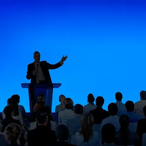 Man in suit speaking at a podium, gesturing to an audience in a room with a blue background.