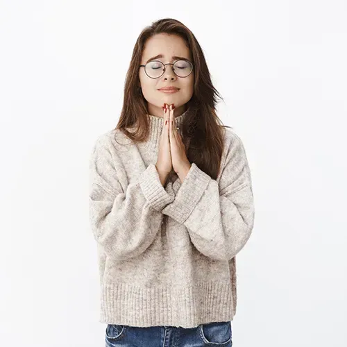 Woman with glasses, hands clasped in front, eyes closed, wearing a sweater and jeans, looking peaceful.
