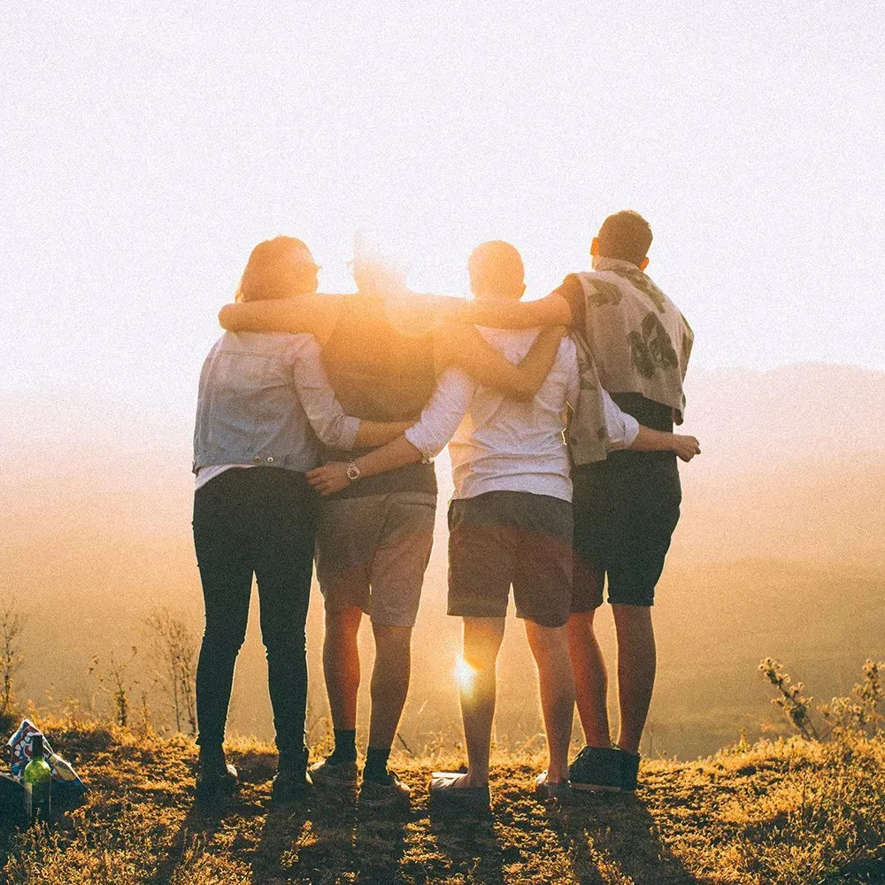 Four friends with arms around each other, looking at a sunset from a hilltop.