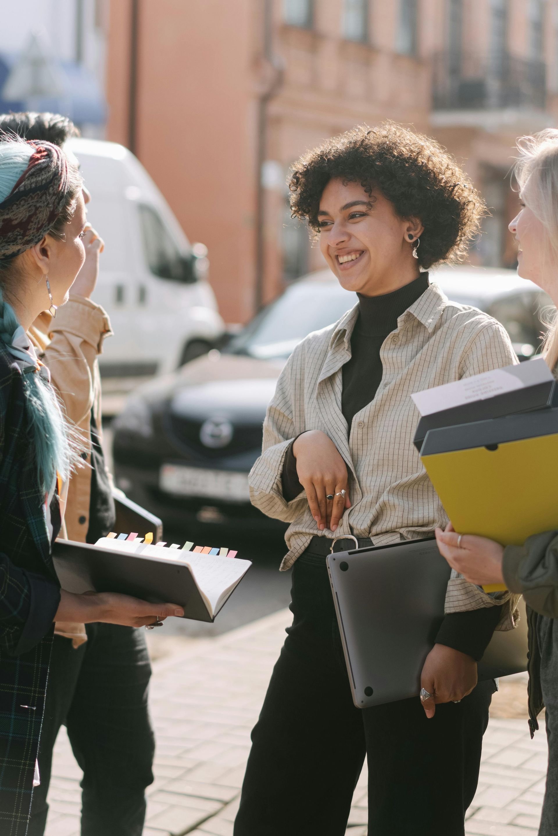 Four people standing outside, talking. Woman in center smiles, holding a laptop. Cars and buildings in the background.