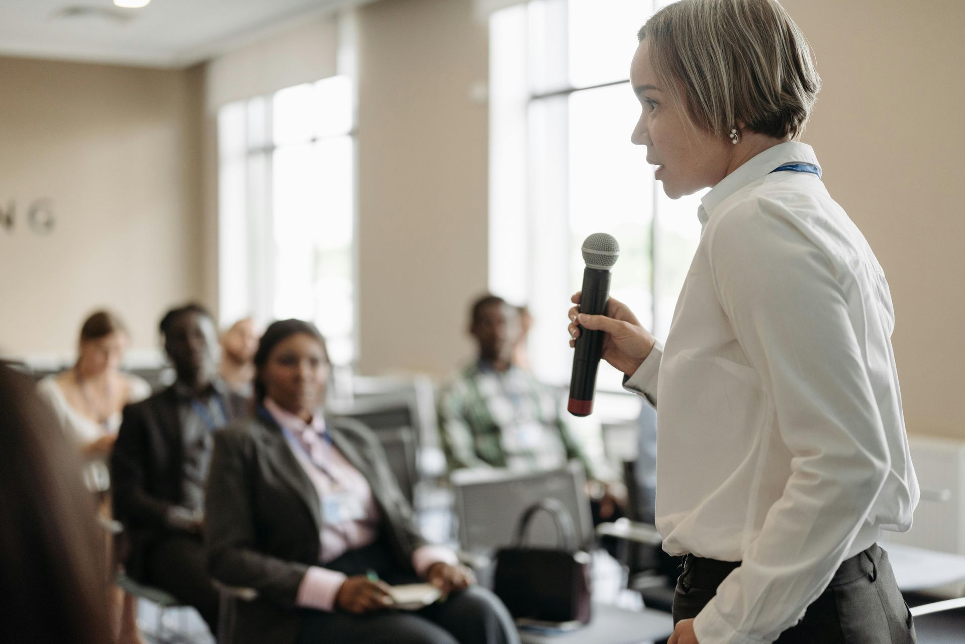 Woman in white shirt speaks into microphone at conference. Audience listens.