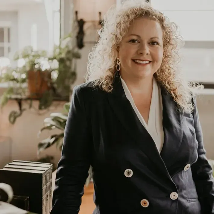 Woman with curly blonde hair smiling, wearing a navy blazer, in a well-lit room with plants.