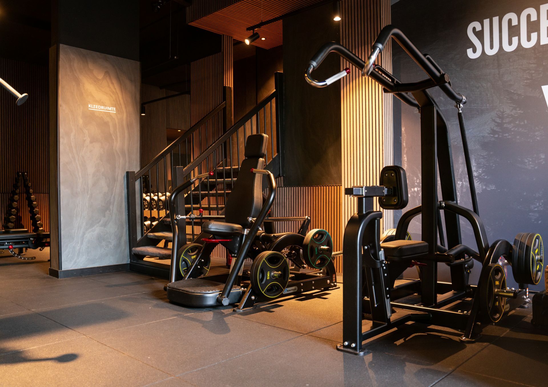 Gewichtsschijven op een halter rusten op de vloer van een sportzaal. Touwen, muren en een emmer op de achtergrond.