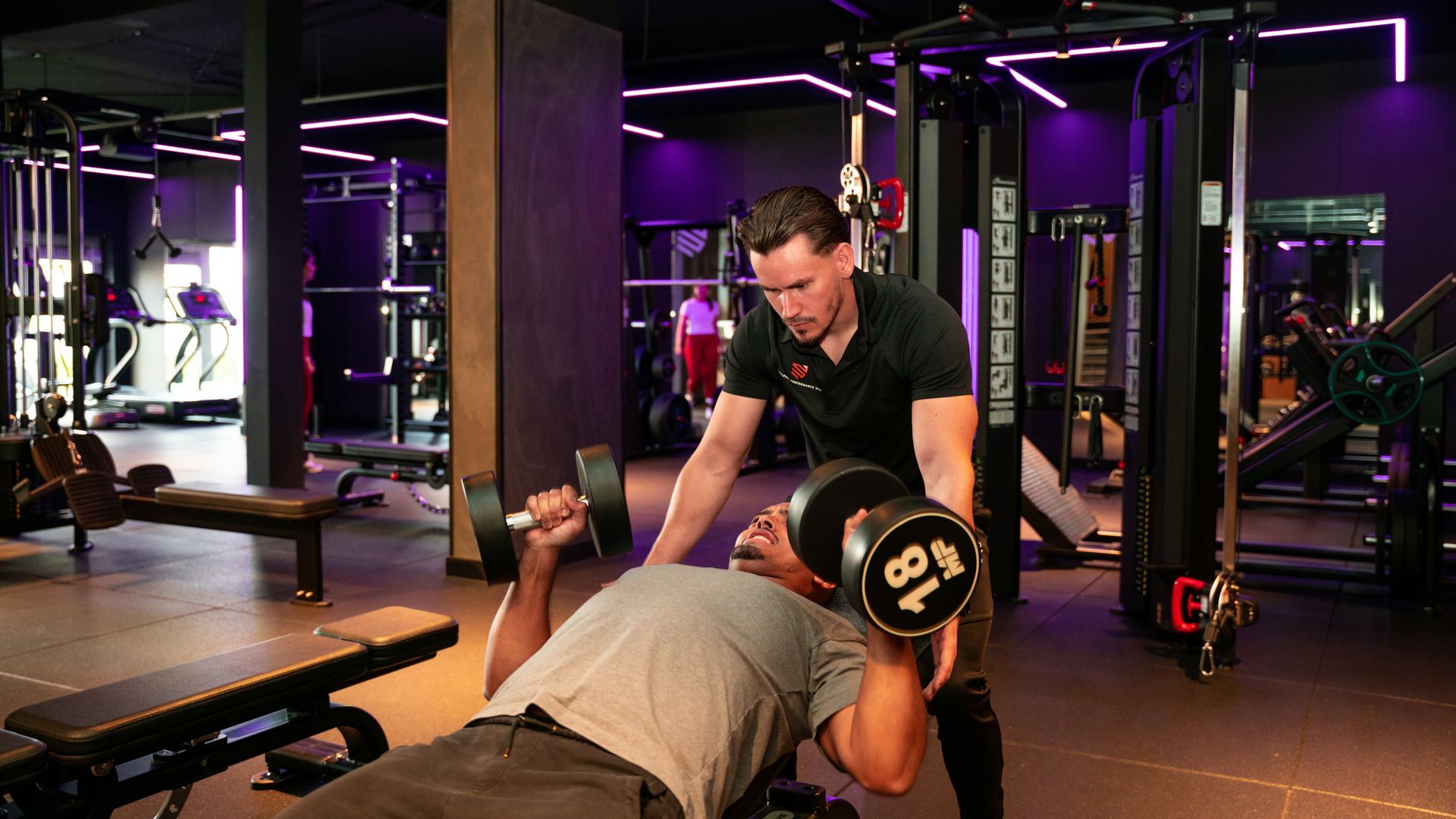 Vrouw tilt een halter op met hulp van een trainer. Sportschoolomgeving.