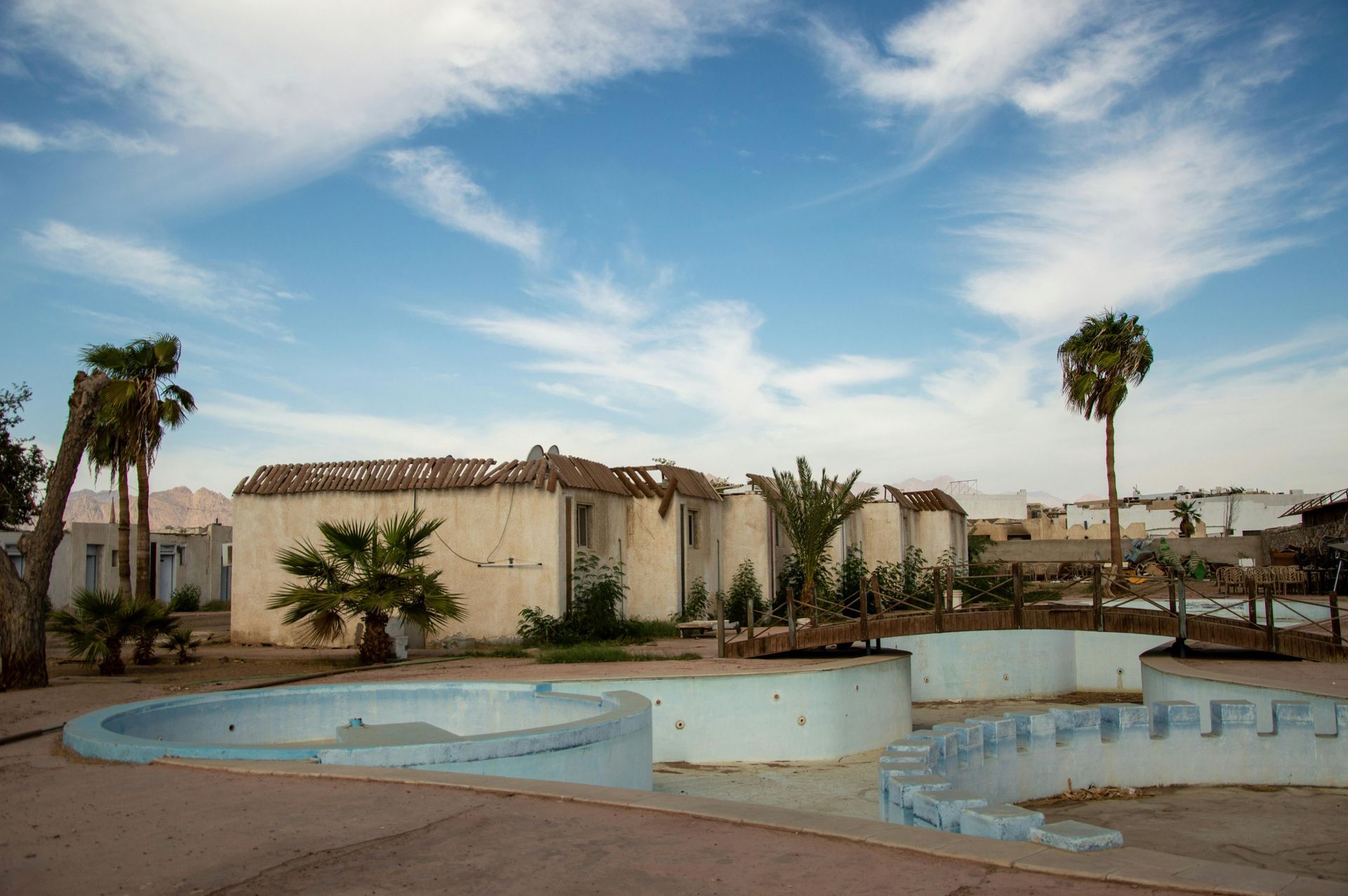 Abandoned buildings with a small pool under a cloudy blue sky. Palm trees stand nearby.