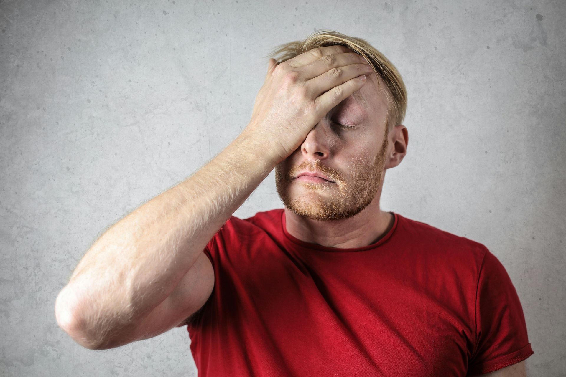 Man in red shirt, hand on forehead, eyes closed, looking stressed against a textured wall.