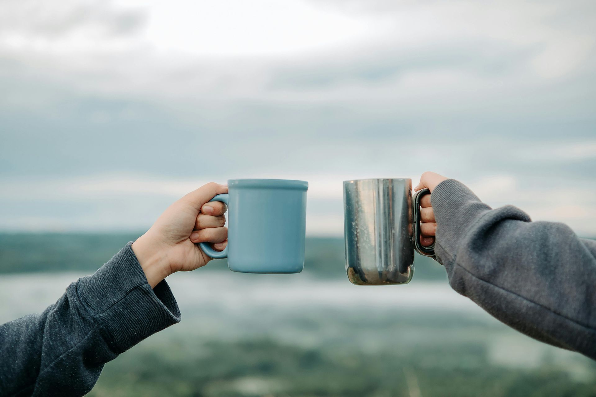 Two people holding up mugs, toasting outdoors with a cloudy sky backdrop and water in the distance.