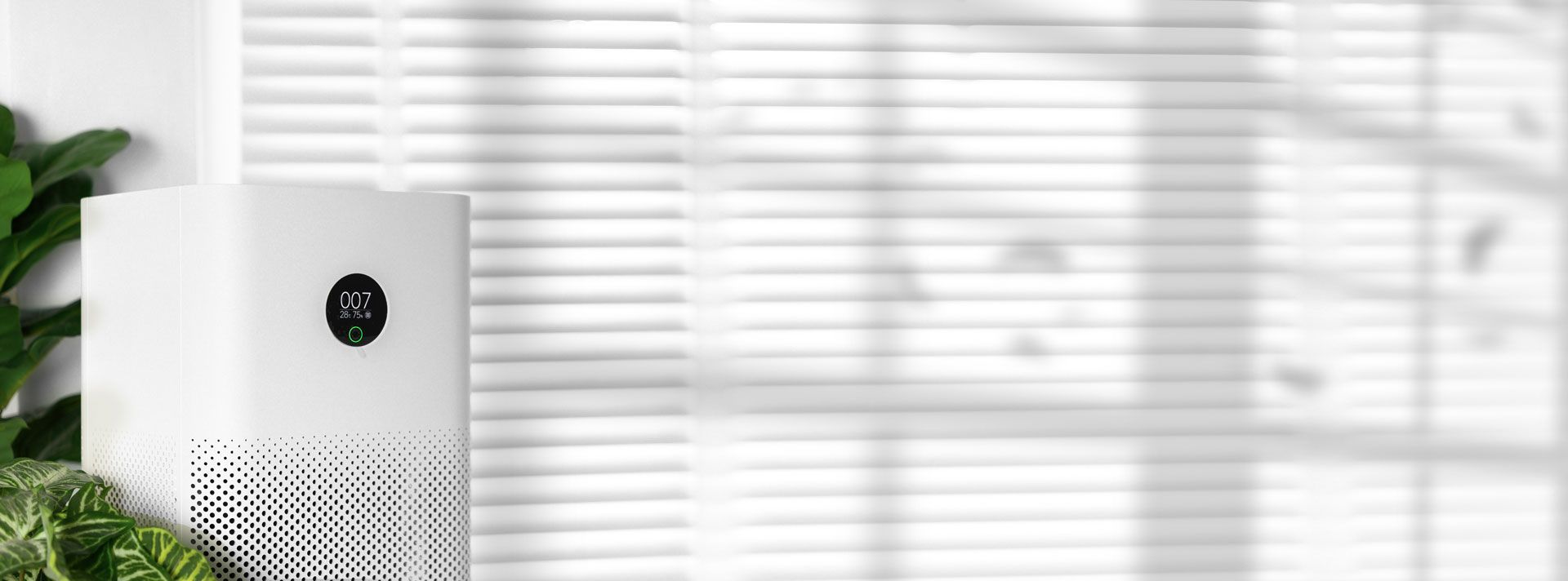 A white air purifier is sitting in front of a window with blinds.