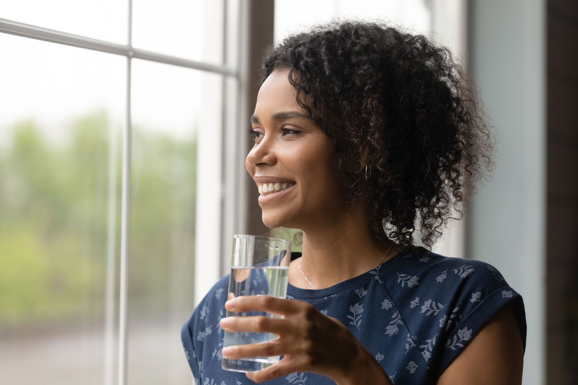 A woman is holding a glass of water and looking out of a window.