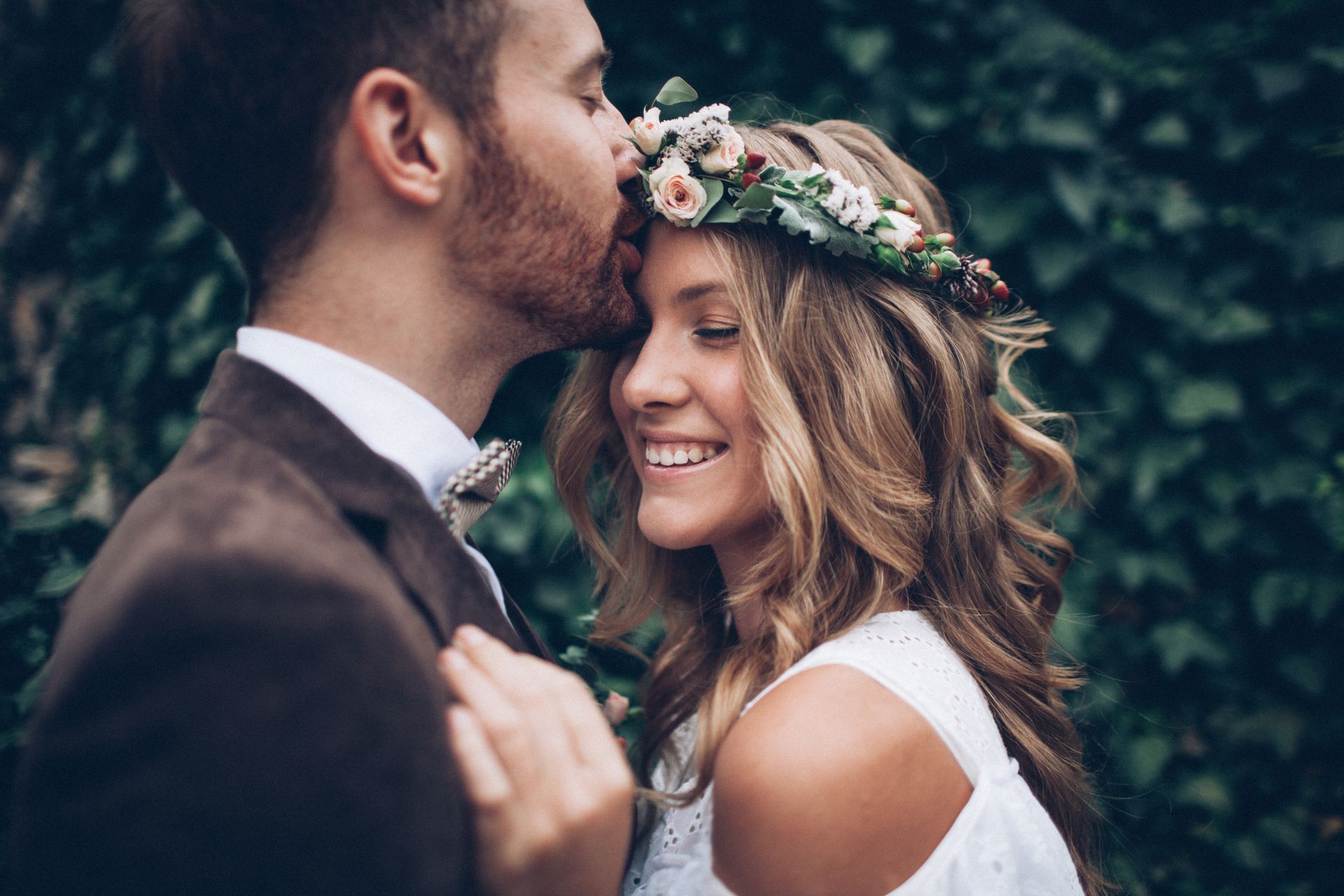 The bride is wearing a flower crown and the groom is kissing her on the forehead.