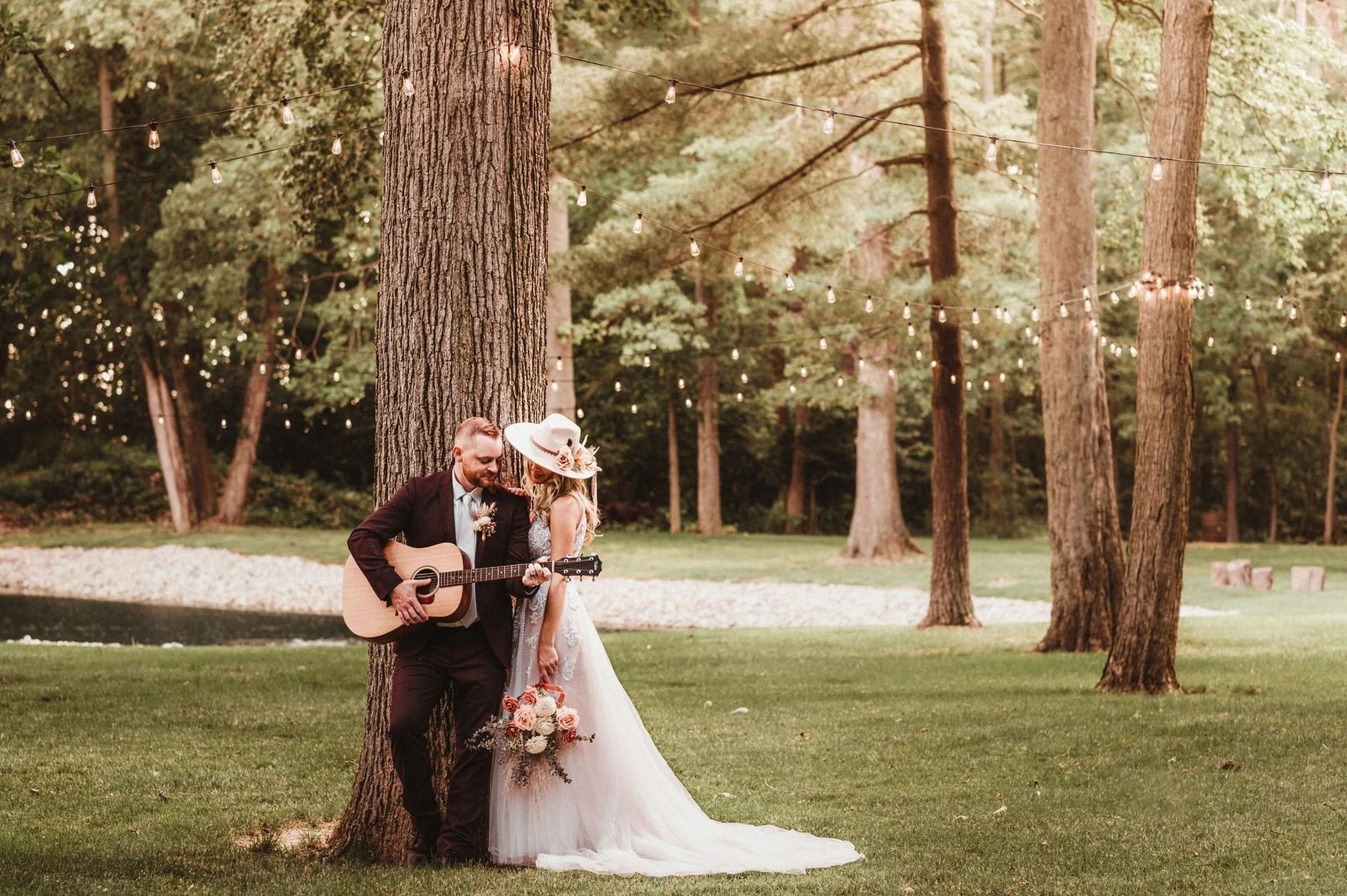 A bride and groom are leaning against a tree while a man plays a guitar.