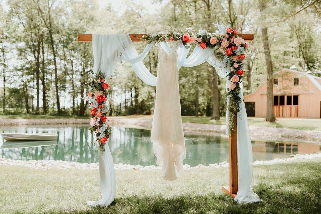 A wedding dress is hanging on a wooden arch in front of a pond.
