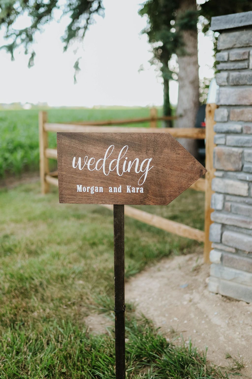 A wooden wedding sign is sitting in the grass next to a stone wall.