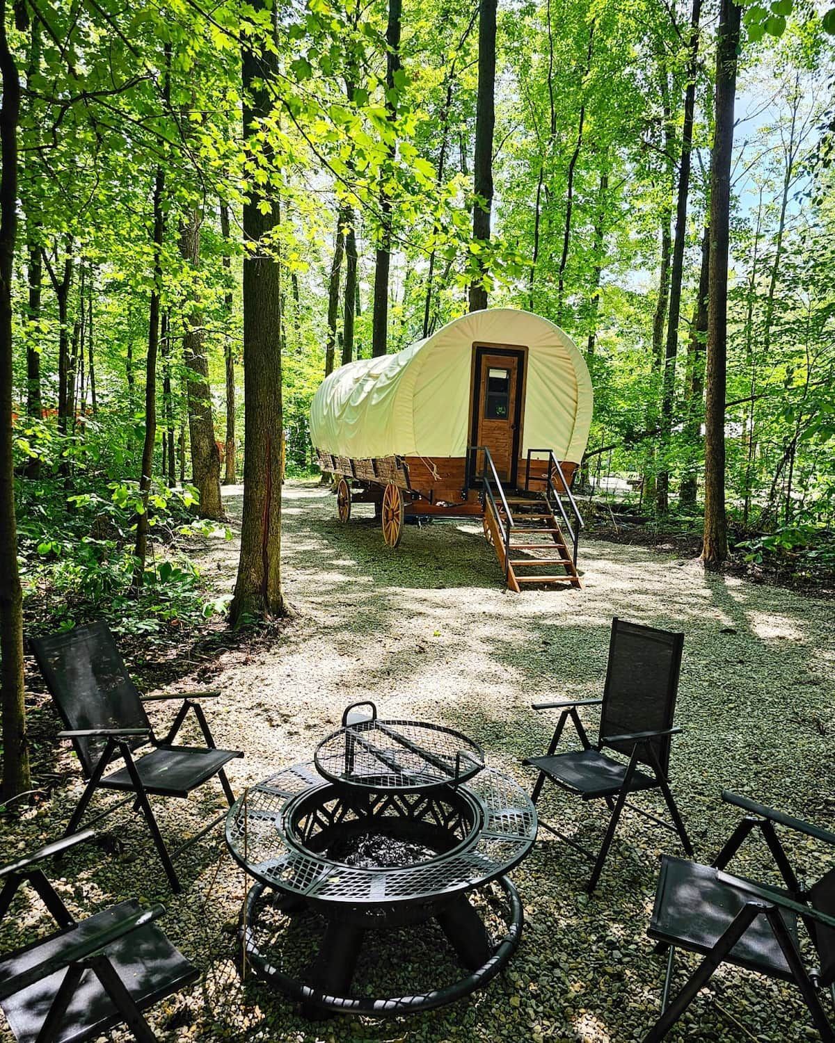 A covered wagon is parked in the woods next to a fire pit.