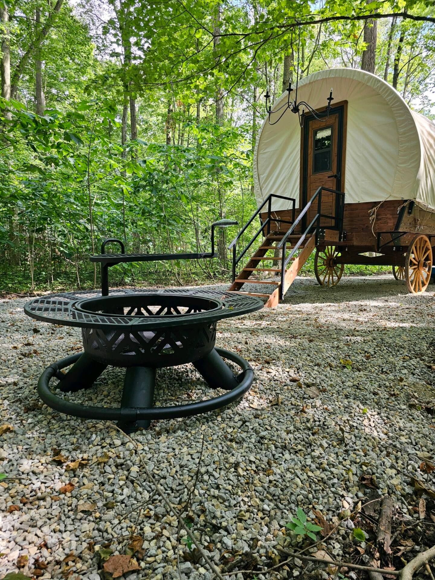 A fire pit is sitting in front of a covered wagon in the woods.