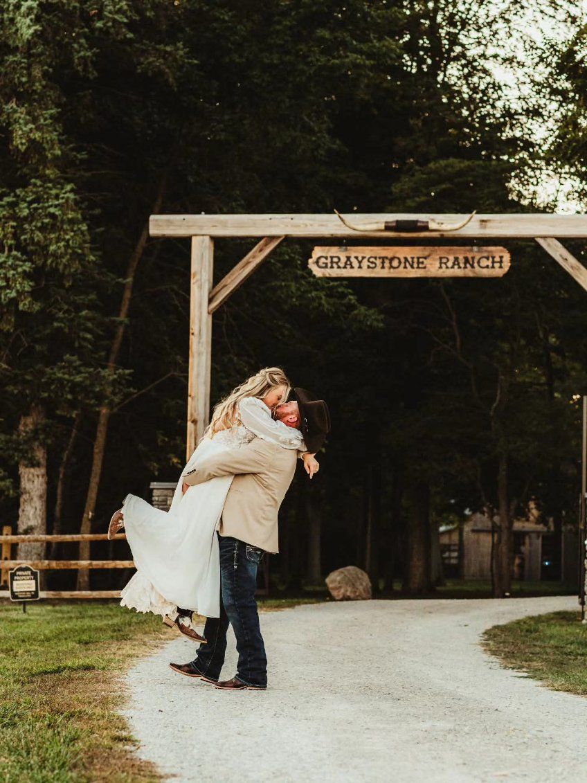 Couple embracing on a gravel path under the Graystone Ranch entrance sign in a wooded setting