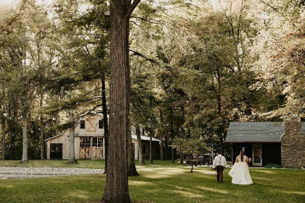 Couple in wedding attire walking across a grassy yard by a rustic house and barn under tall trees