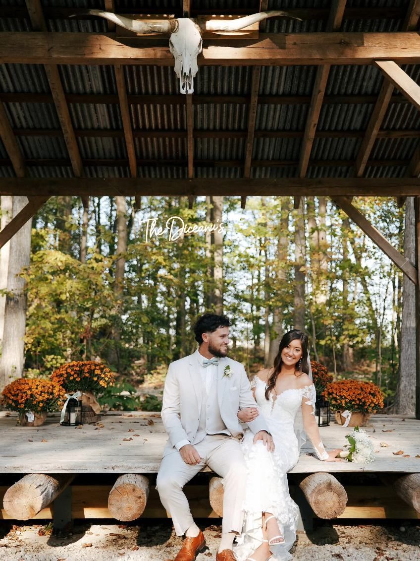 Couple in white wedding attire seated under a rustic pavilion with flowers and trees in the background