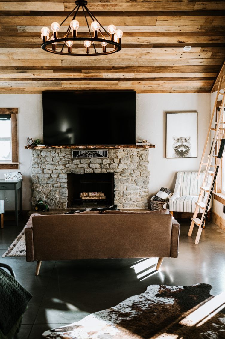 Rustic living room with stone fireplace, mounted TV, wood ceiling, and brown sofa