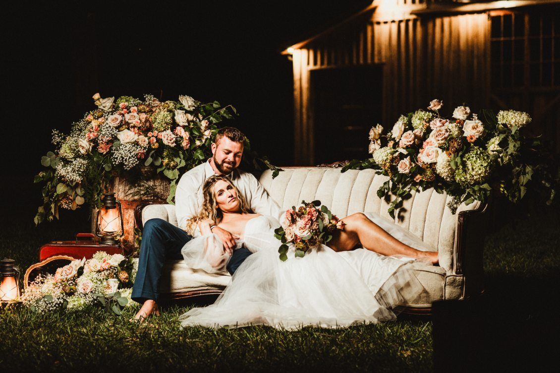 Couple in formal attire reclining on a couch at a floral outdoor wedding reception at night