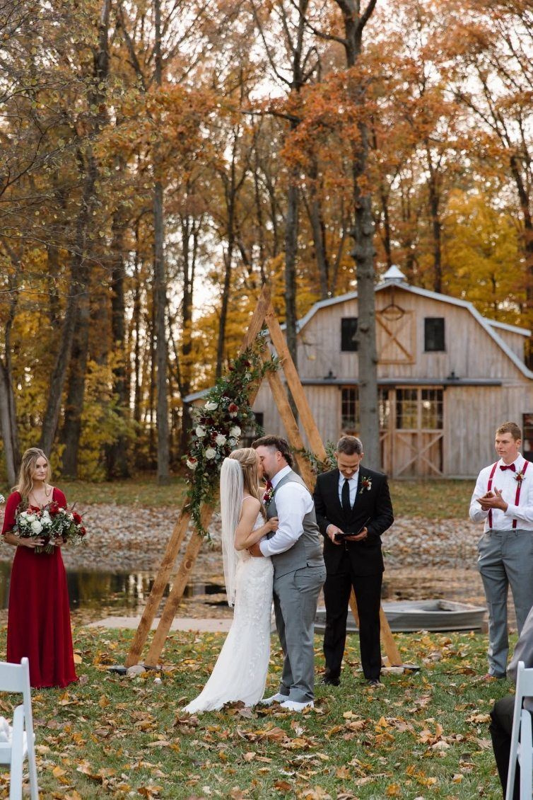 Bride and groom kissing outdoors at a rustic autumn wedding ceremony, with guests nearby and a barn in back