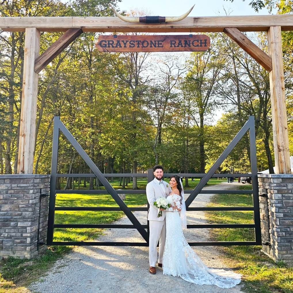Newlywed couple in white standing at the Graystone Ranch gate, holding a bouquet outdoors