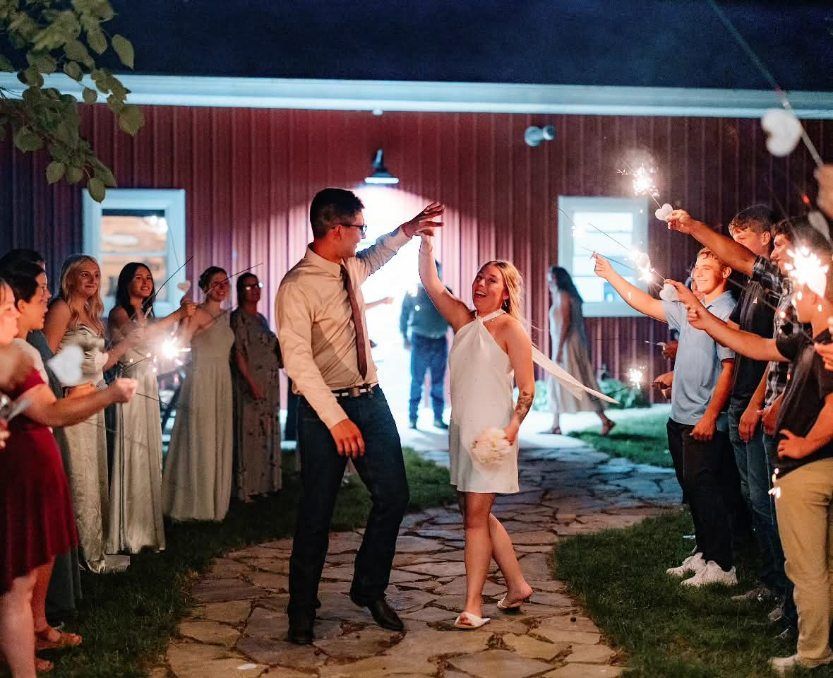 Wedding guests line a stone path with sparklers as a couple walks between them outside a red barn at night