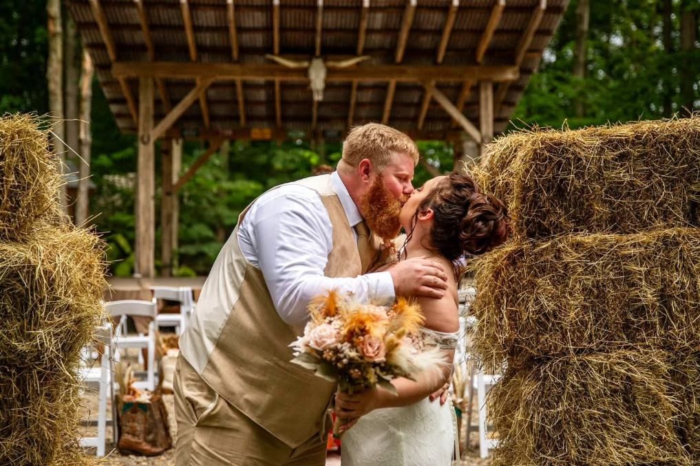 Couple kissing in a rustic barn aisle, surrounded by hay bales and wedding decor.