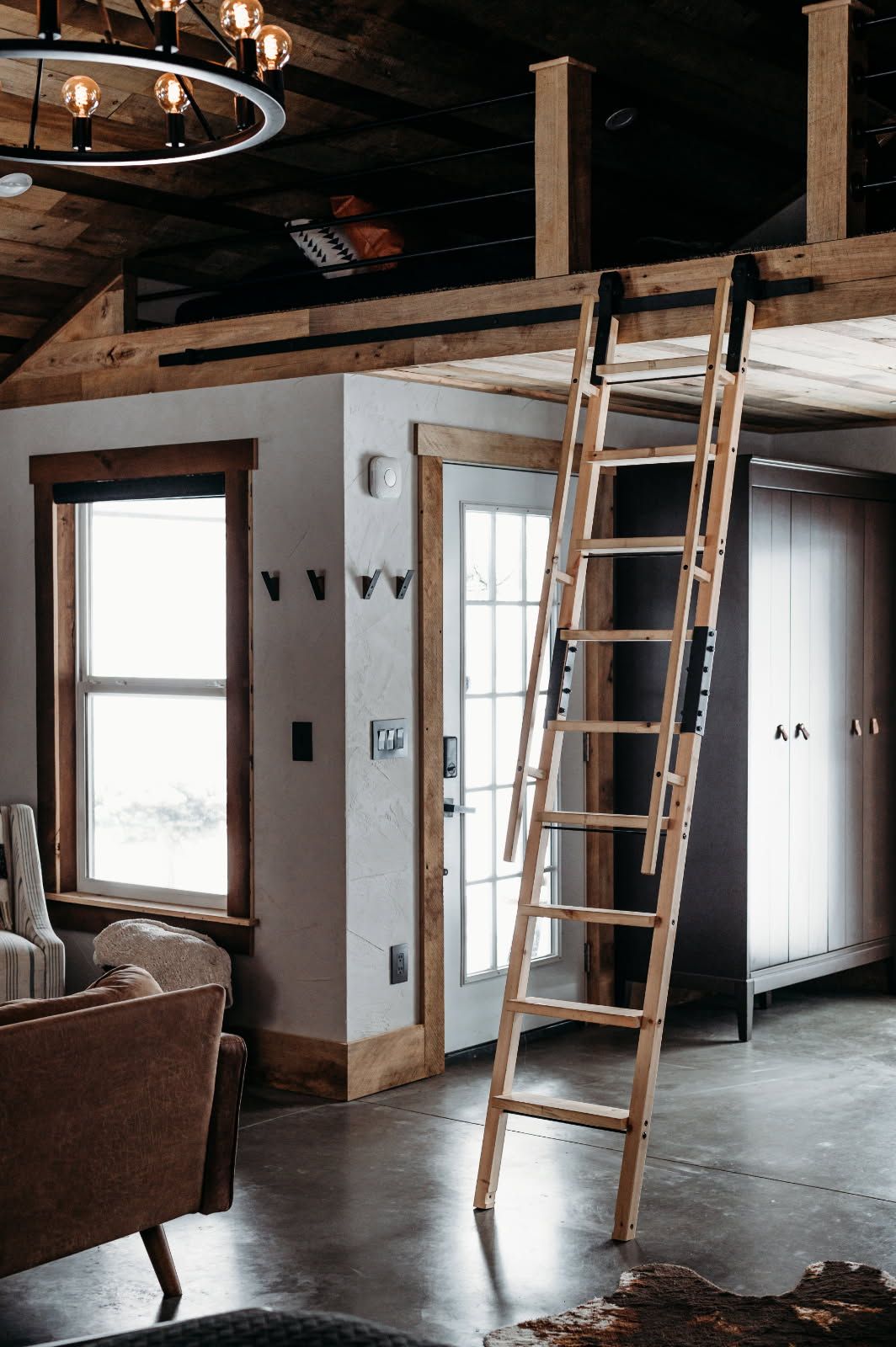 Loft interior with a wooden ladder leading to a mezzanine, exposed beams, and windows with white walls