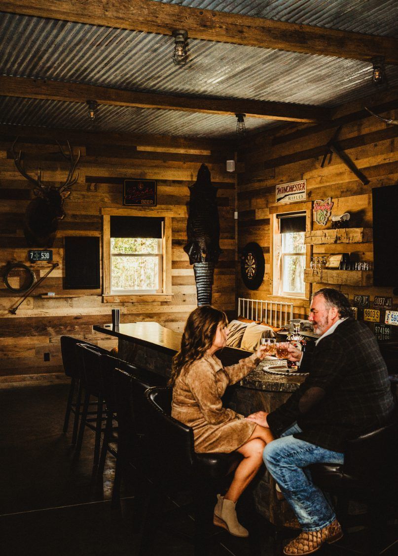 Two people talking at a rustic bar in a cozy wooden cabin, warm light and shelves in the background
