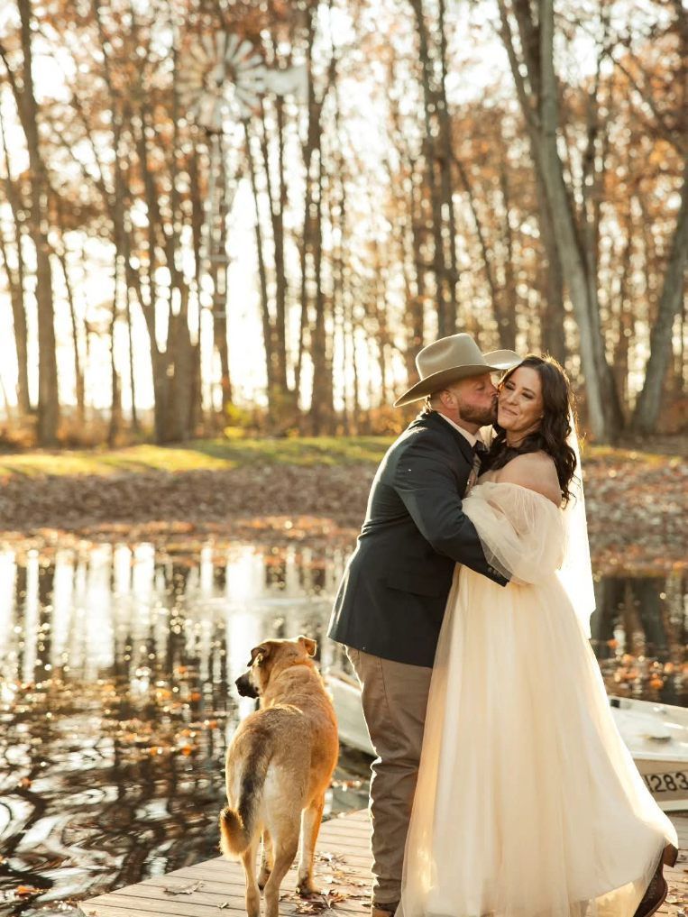 Couple embracing by a pond at sunset, with a dog standing nearby in a wooded park