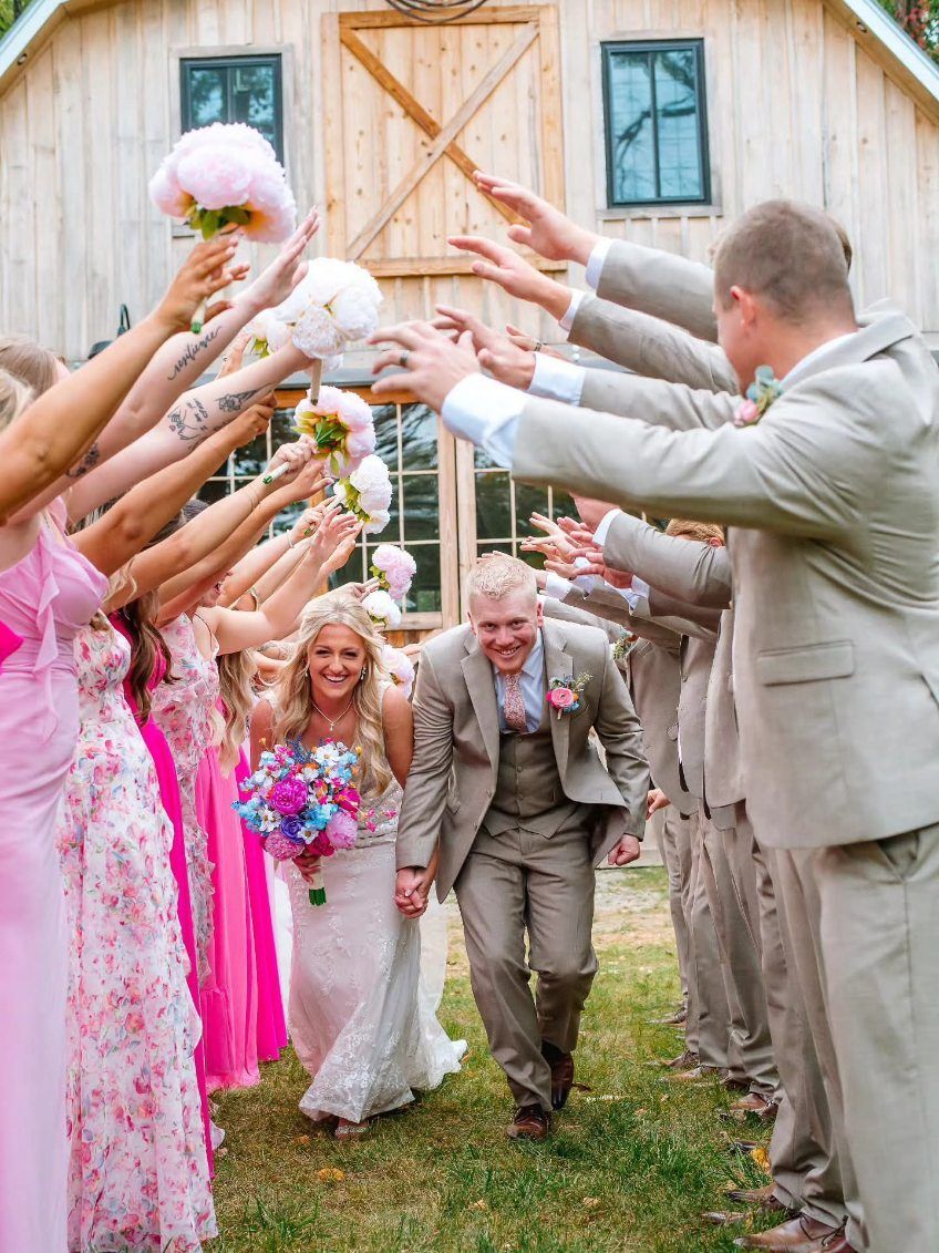 Wedding couple walking under a floral arch of raised hands outside a rustic barn